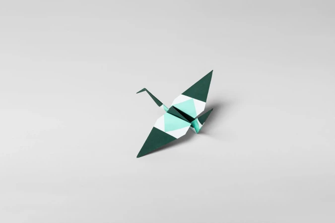 Close-up of a paper crane made of folded paper in teal and dark green, centered on a plain white background. Paper art.