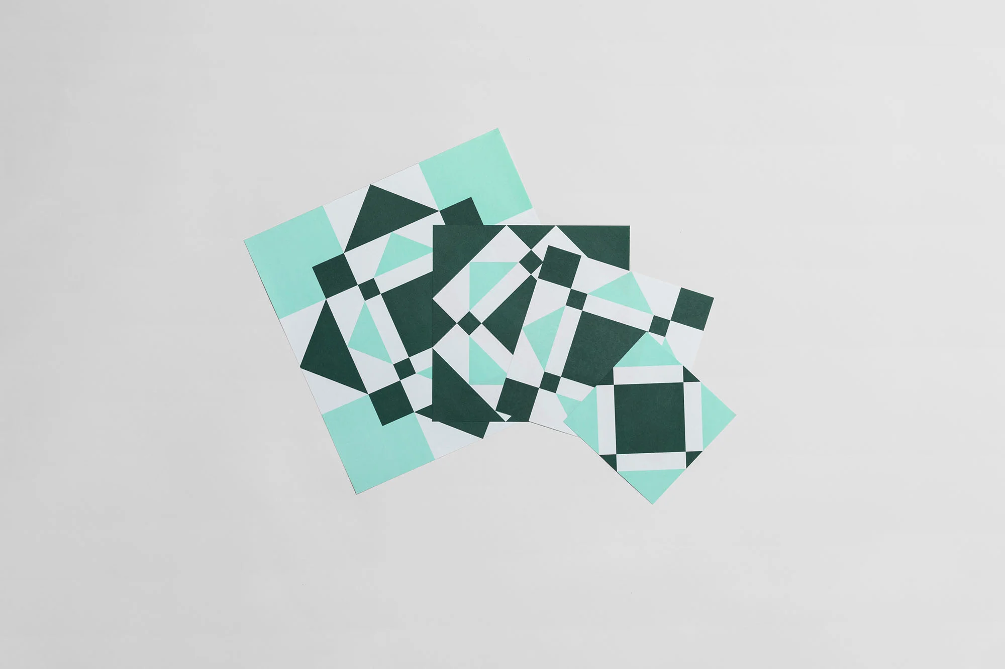 Geometric paper art: Three overlapping square papers with graphic patterns in teal, forest green, and white on a light gray background.