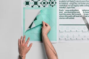 Close-up of hands peeling back a turquoise and white patterned paper print on a grey surface with metal ruler and craft knife.
