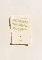 Close-up shot of a textured paper card with printed olive oil brand story and green logo against a neutral background