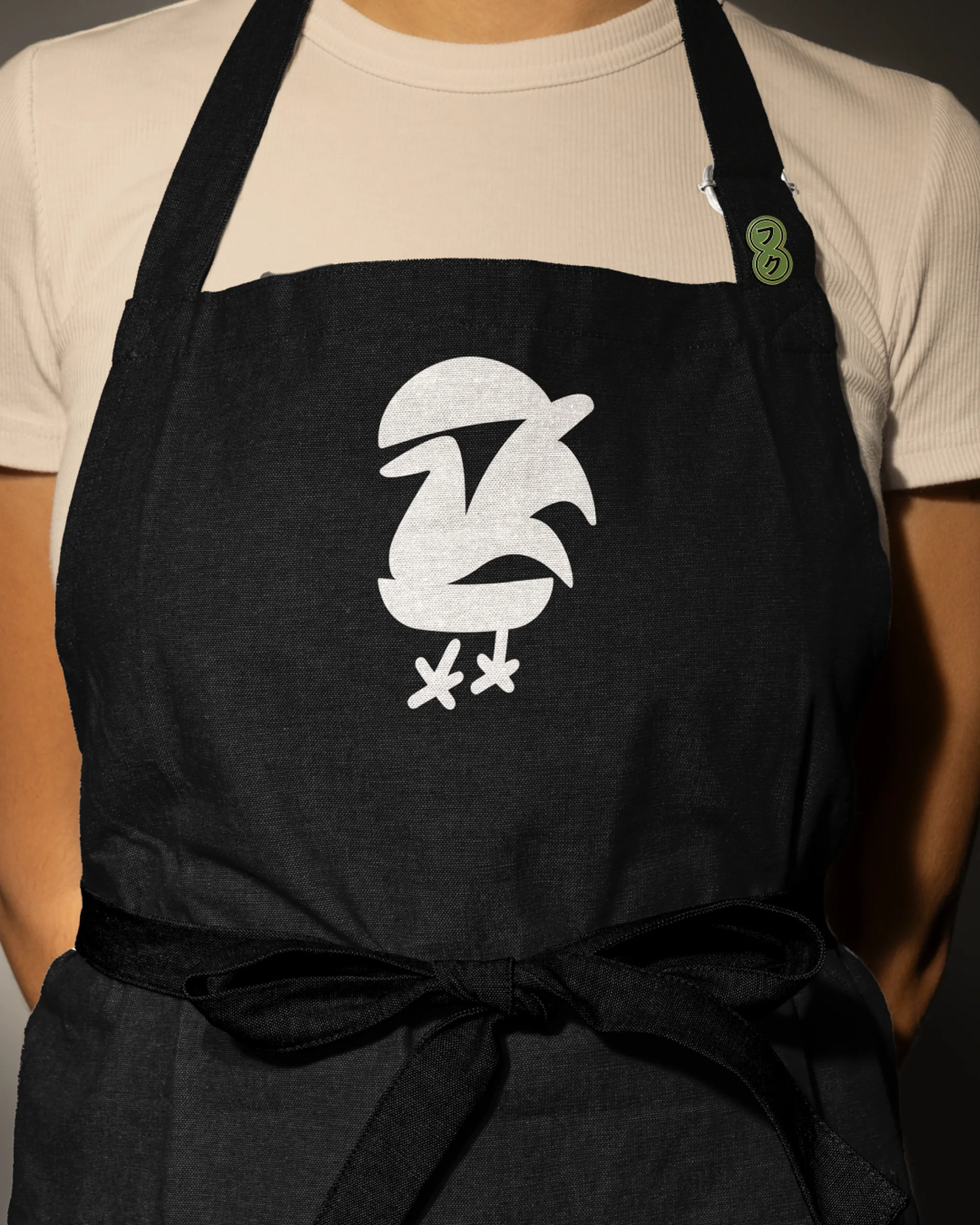 A person wears a black apron with a unique white bird graphic and tied waist, against a neutral background, in a close-up studio shot.
