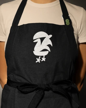 A person wears a black apron with a unique white bird graphic and tied waist, against a neutral background, in a close-up studio shot.