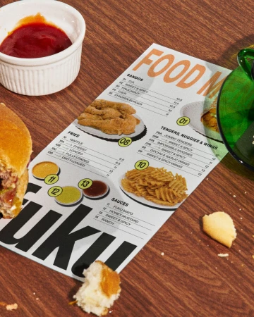 Overhead shot of a menu featuring food items like a burger, chicken tenders, fries, and sauces on a wood surface with food debris.