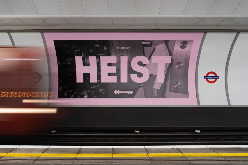 Illuminated advertising poster in a subway featuring the word HEIST in large pink letters against a background of dark machinery.