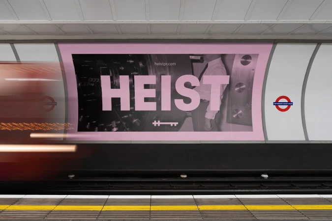 Illuminated advertising poster in a subway featuring the word HEIST in large pink letters against a background of dark machinery.
