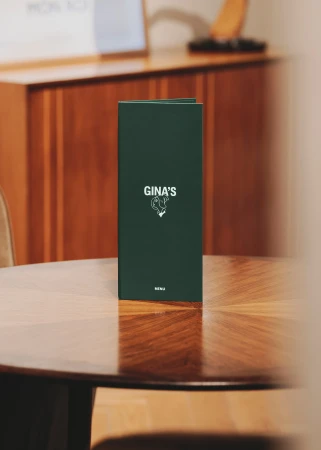 Menu on a round wooden table in a restaurant setting, featuring the name 'Gina's' on its cover with a simple illustration.