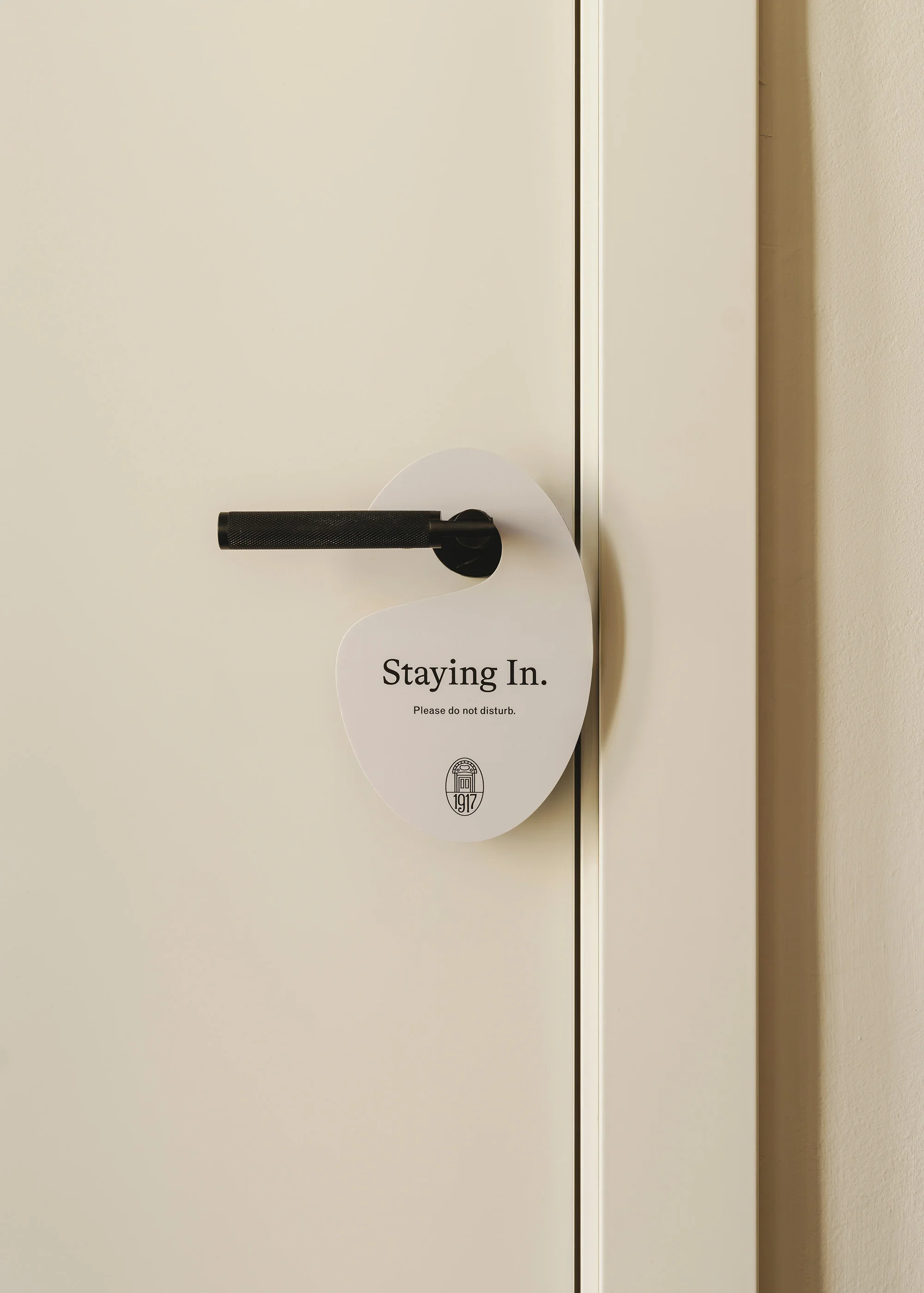 Door hanger on a white door with a black handle, displaying the text 'Staying In. Please do not disturb.' in black font.