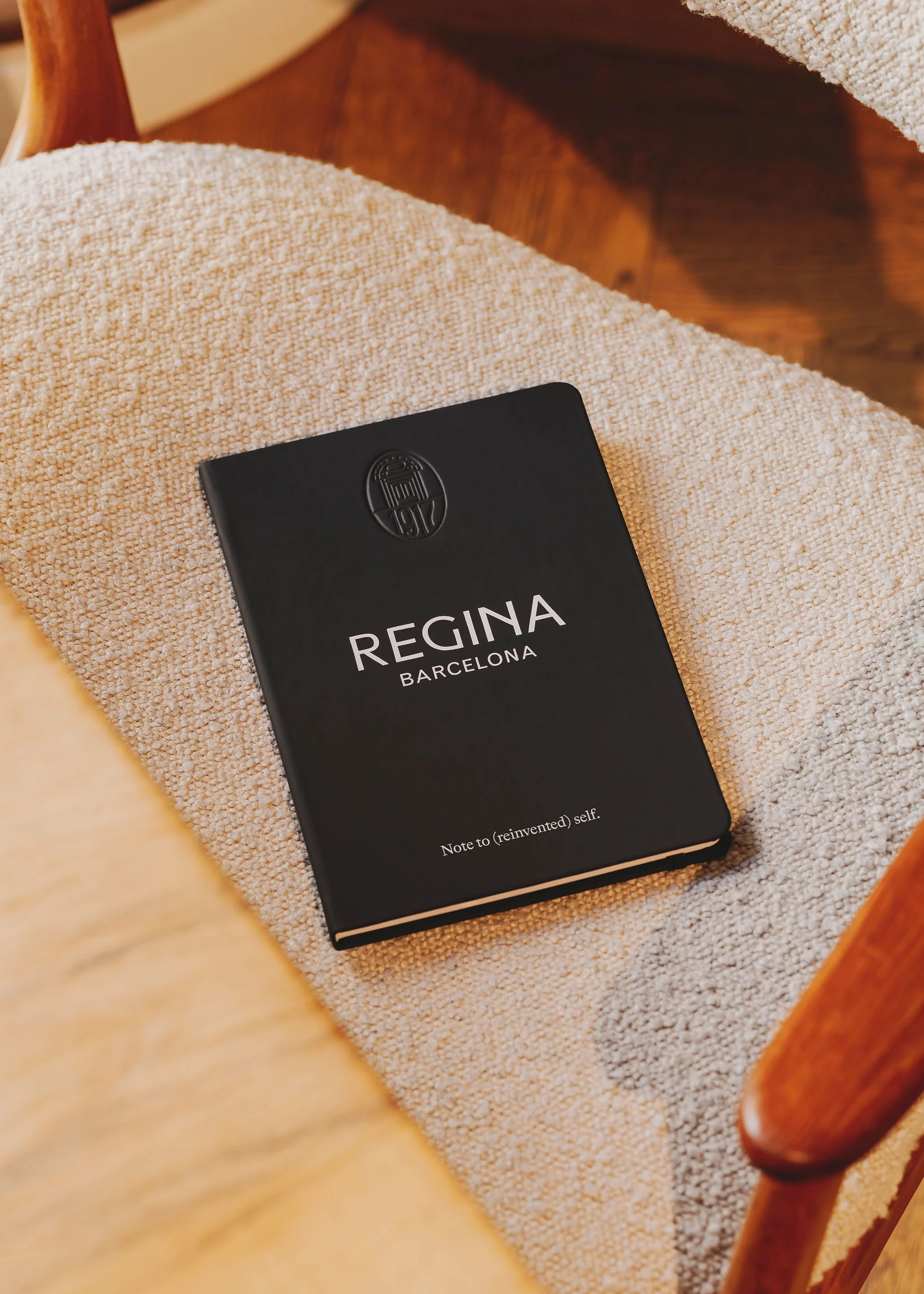 Black notebook with 'Regina Barcelona' on a white, textured chair with wooden armrests in soft lighting, creating a simple, elegant composition.