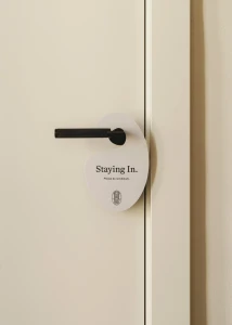 Close-up of a white organic shaped door hanger reading Staying In on a cream door with a black handle for Hotel Regina brand identity.
