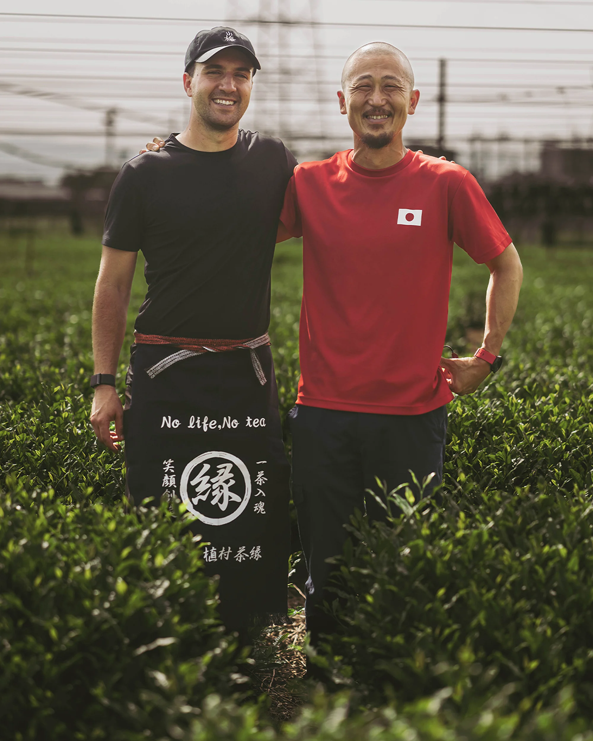 Two men smile in a tea field; one wears an apron with Japanese text, and the other a Japanese flag shirt, highlighting cultural exchange.