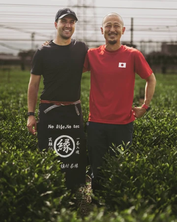 Two men smile in a tea field; one wears an apron with Japanese text, and the other a Japanese flag shirt, highlighting cultural exchange.