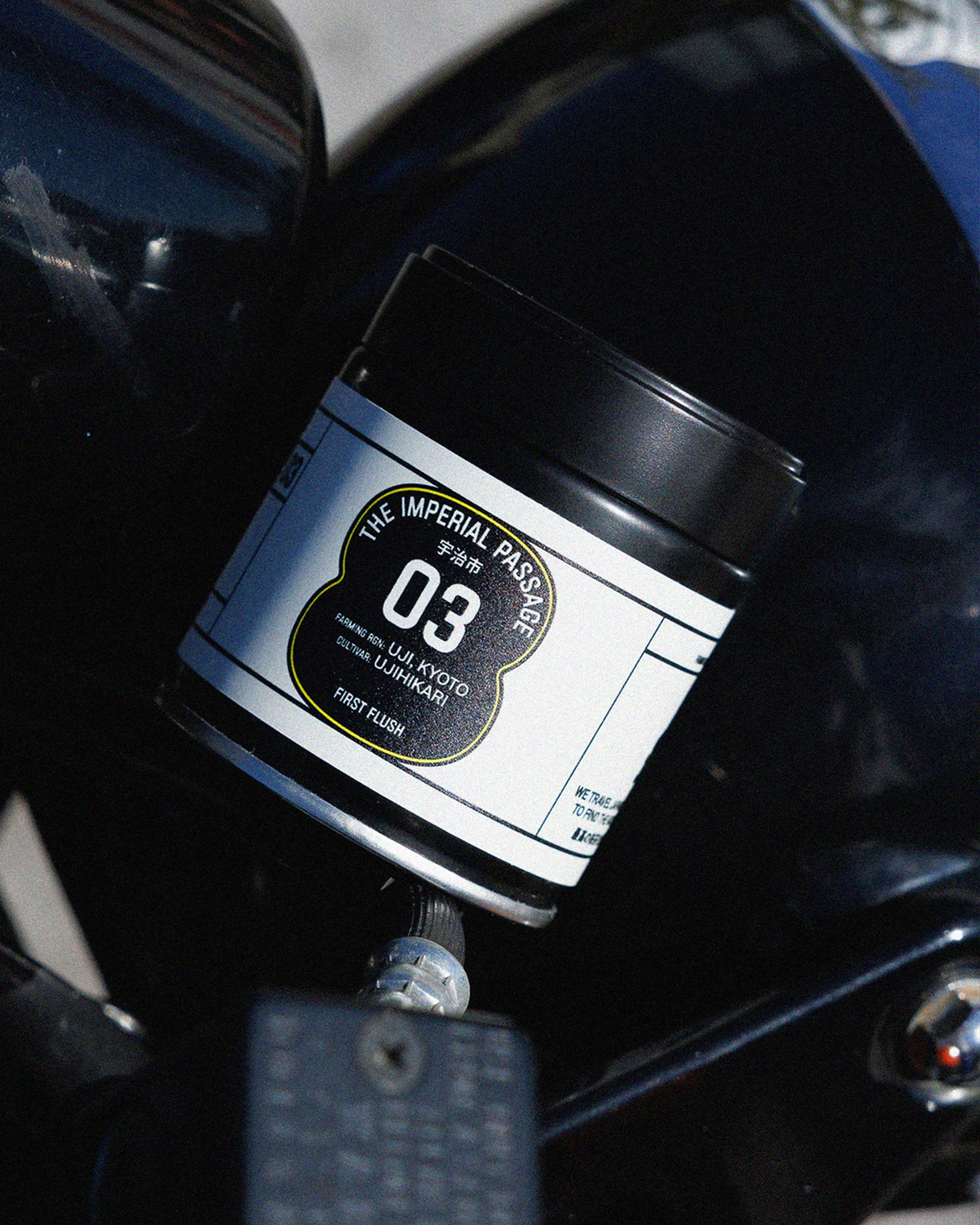 Close-up of black and white product container with Japanese text and 'The Imperial Passage 03' on a black motorcycle.