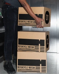 Person holding a branded brown cardboard box over a stack of two matching boxes for Motorcycle Matcha
