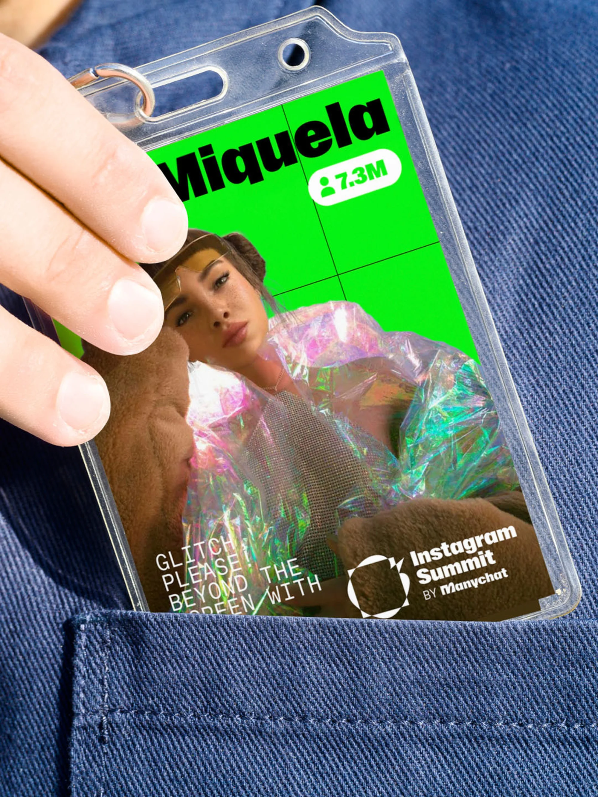 Hand holding a name badge with a woman's photo on a bright green background. The badge is tucked into a blue denim pocket.