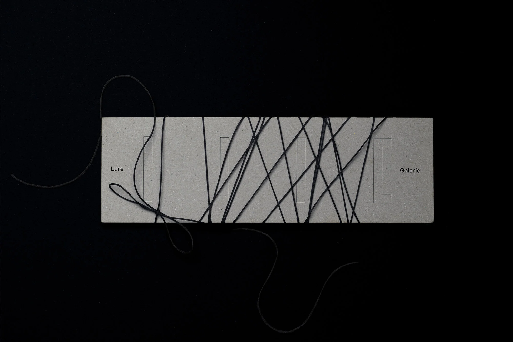 Overhead shot of a grey rectangular box with black strings, featuring the words 'Lure' and 'Galerie' on a black background.