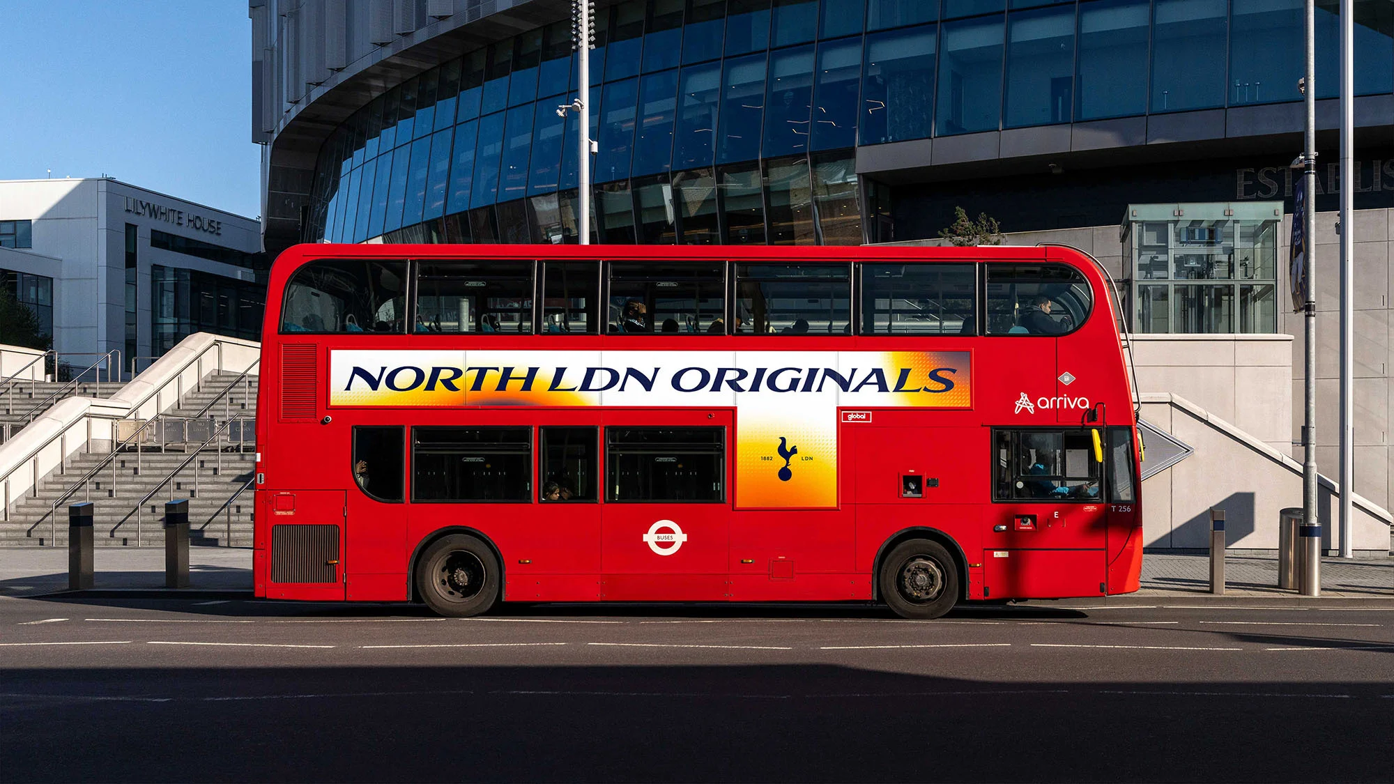 A red double-decker bus with 'NORTH LDN ORIGINALS' advertisement is parked in front of the Tottenham Hotspur Stadium, London, England.