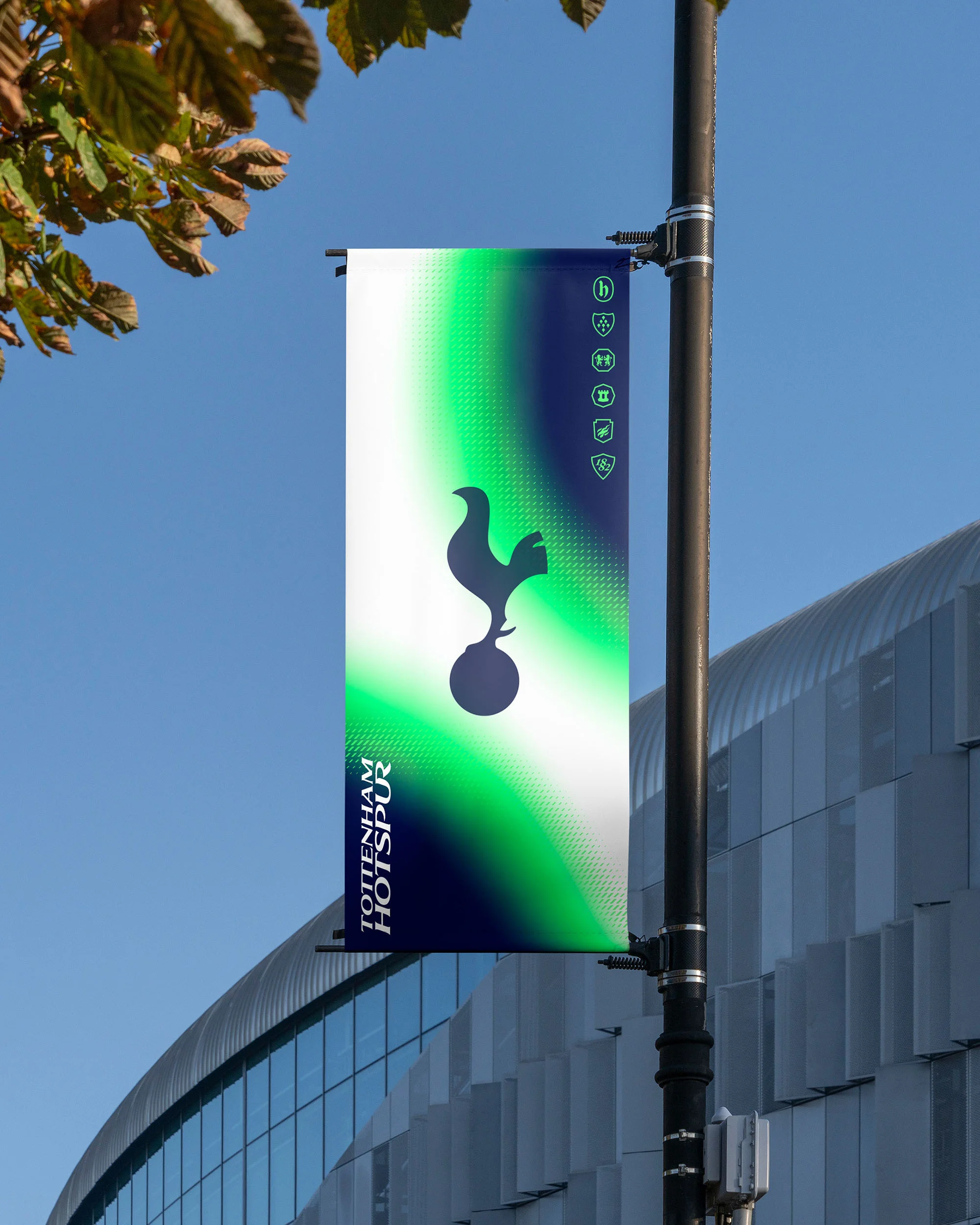 Tottenham Hotspur soccer team banner on a light pole in front of a stadium with blue sky and leaves in the background.