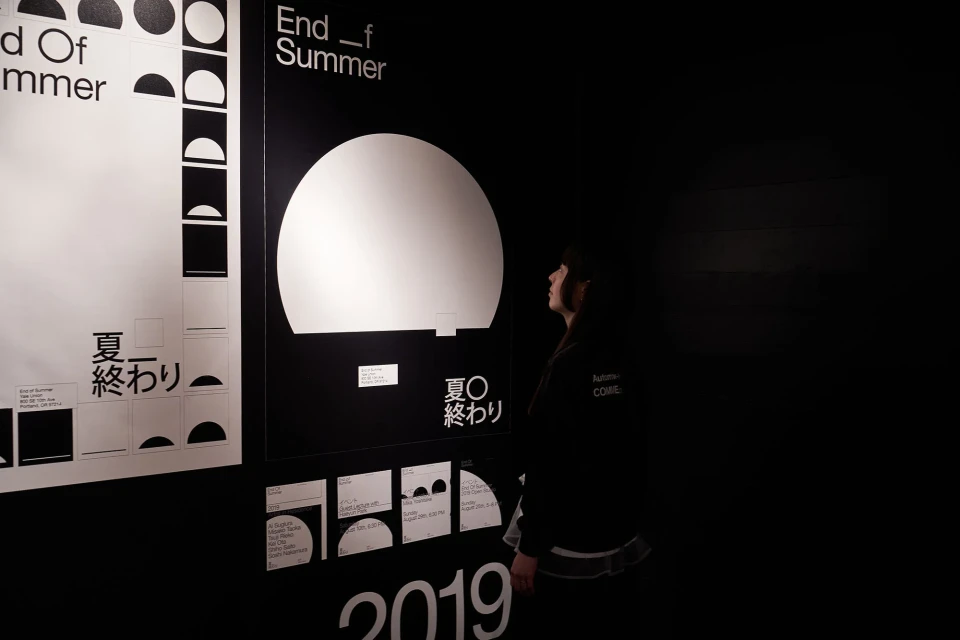 A young woman views black and white graphic design posters in a dark, indoor exhibit space, featuring bold typography and geometric shapes.