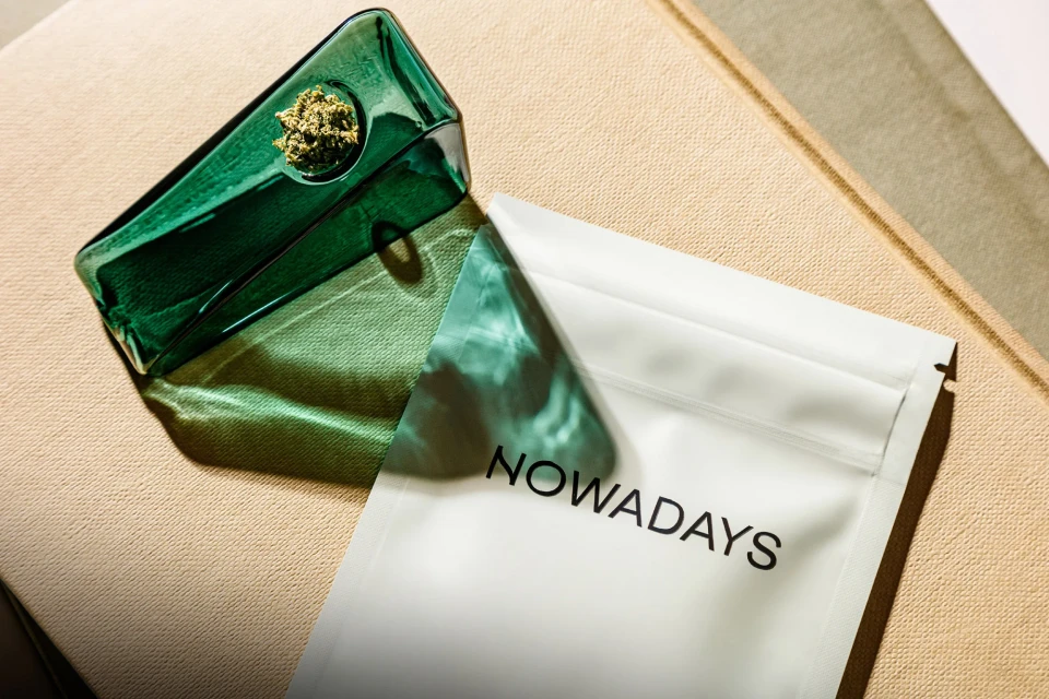 Close-up of a green glass pipe filled with cannabis next to a white package with text 'NOWADAYS' on a textured beige surface.