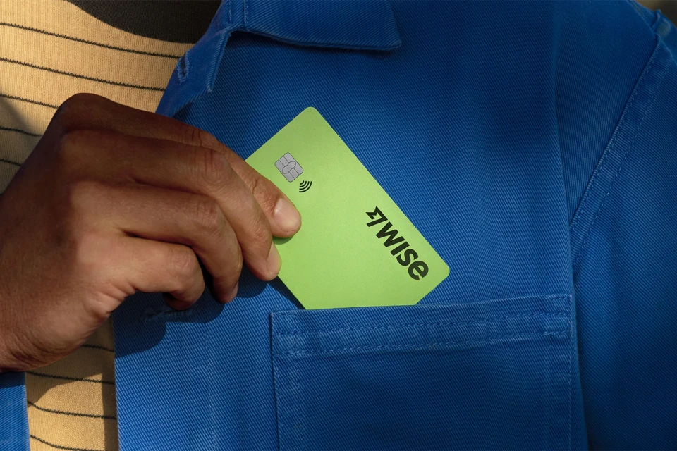 Close-up of a hand placing a light green Wise card into the pocket of a blue shirt against a horizontally striped background.