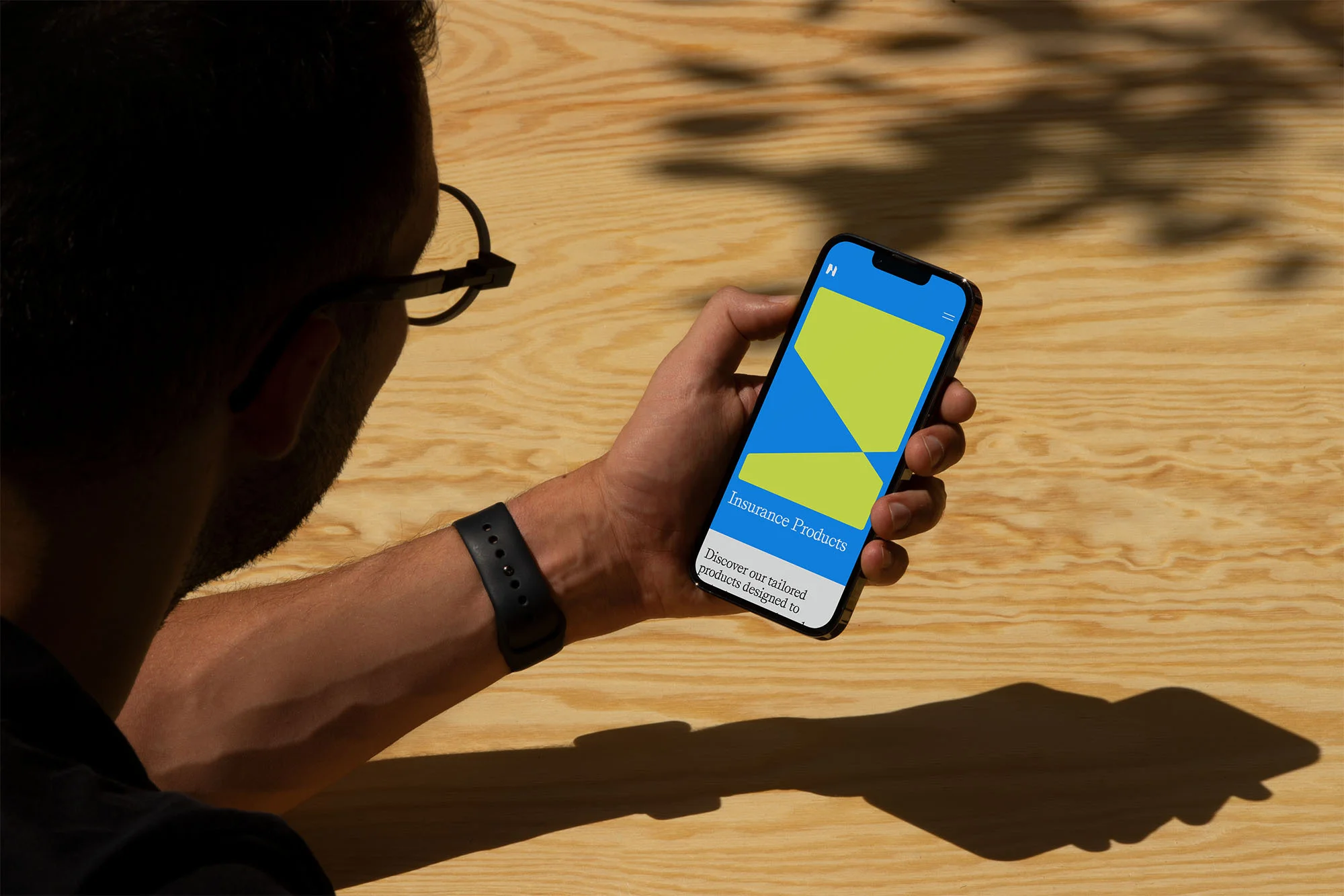 Adult male holds a mobile phone with an insurance app, showcasing blue and green geometric shapes, against a wood background outdoors.