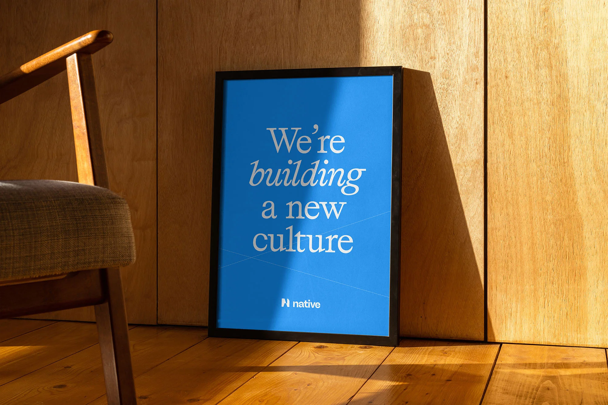 Framed blue poster leaning against a wood wall, featuring the text: We're building a new culture, with modern chair visible.