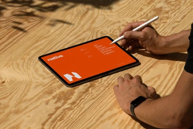 Close-up of a person using a stylus on a tablet displaying the Native brand identity against a textured wooden surface
