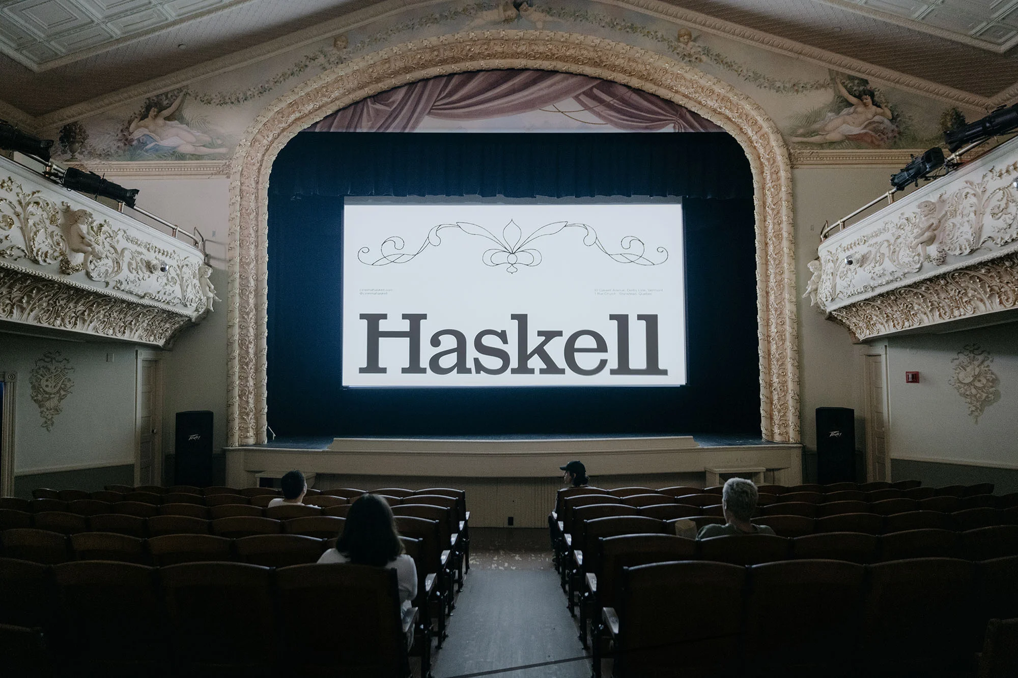 Interior view of a theatre with rows of empty seating facing a stage screen displaying the word 'Haskell' in bold font.