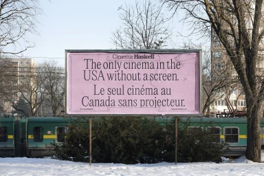 Pink billboard reading Cinema Haskell with bilingual text outdoors in front of a green train and bare winter trees