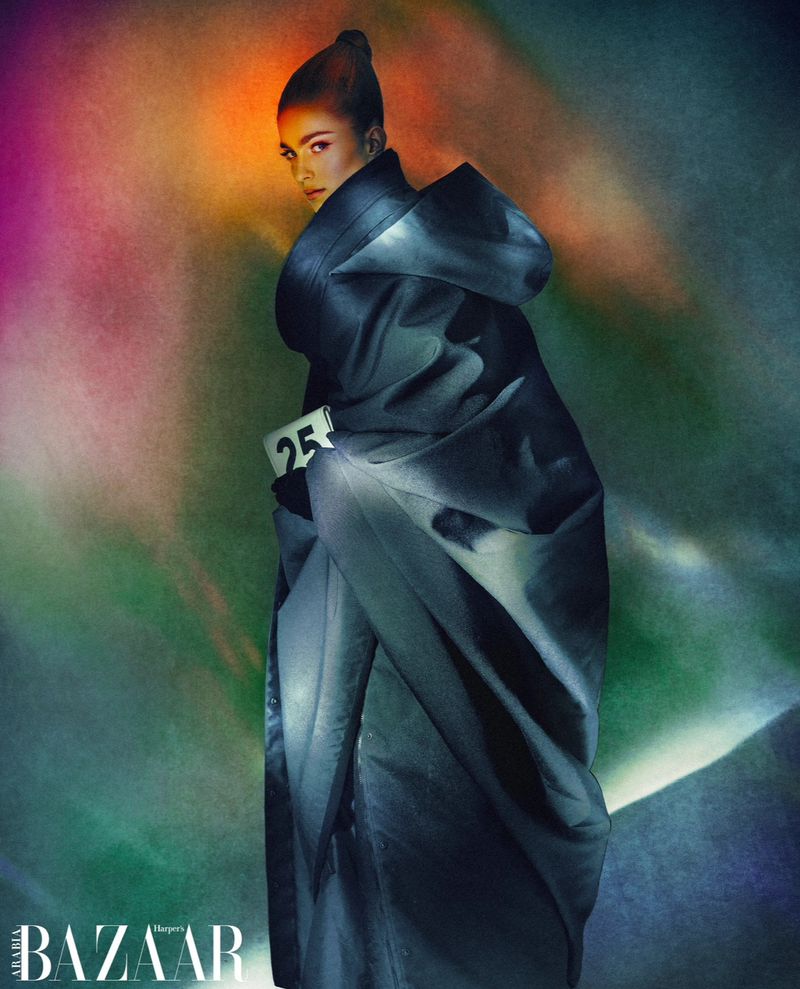 Fashionable woman poses in a draped coat against a blurred abstract backdrop with soft lighting and BAZAAR logo, creating a stylish editorial shot.