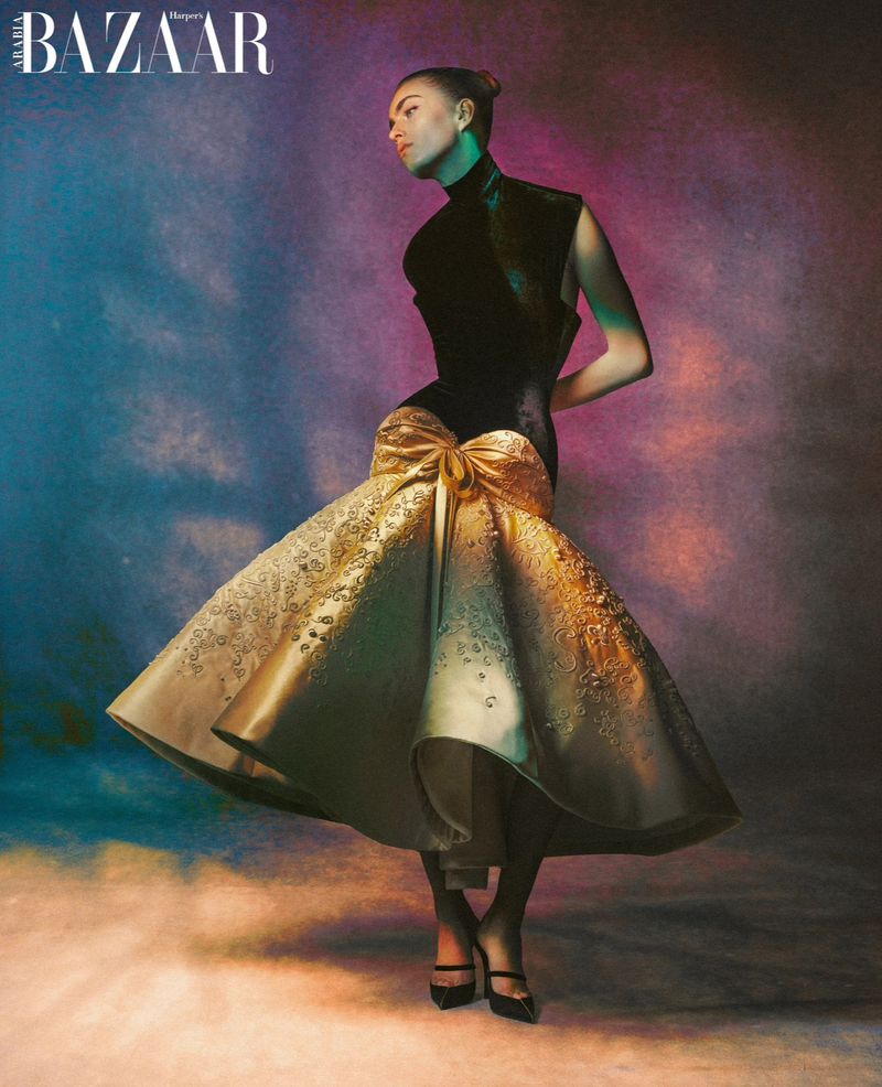Fashion editorial shot of an adult female model in a black sleeveless top and gold skirt with bow, in a studio with colorful lighting.