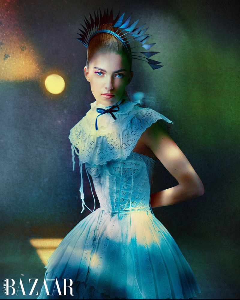 Woman in frilly, blue-tinted dress with headpiece poses in a fashion editorial style image with moody lighting and a textured backdrop.