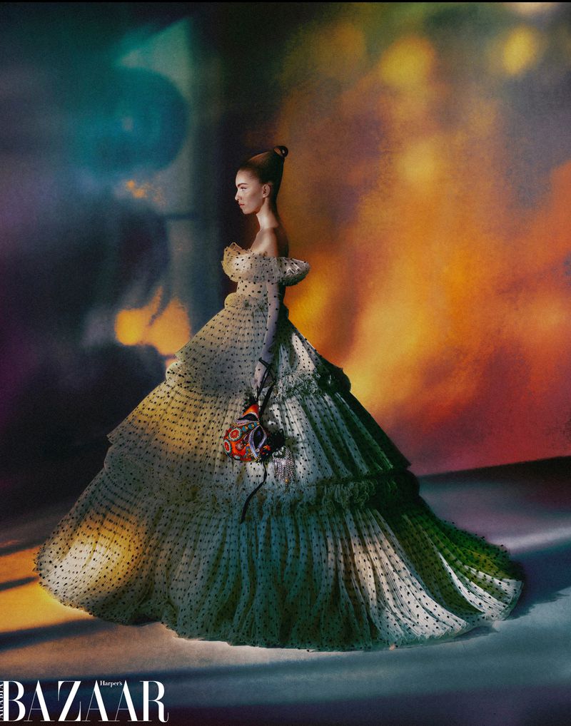 Elegant woman in a formal dress posing against a studio backdrop with a colorful, abstract design and visible "Harper's BAZAAR" logo.