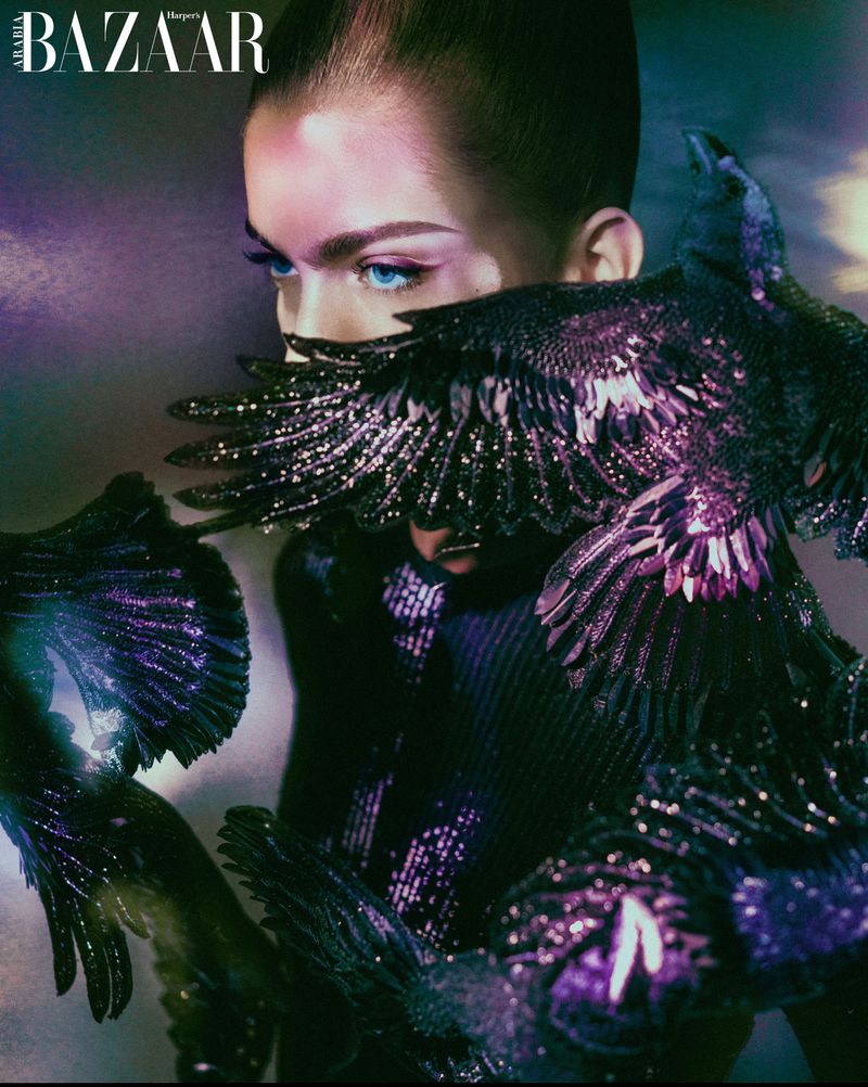 Fashion shot of a model wearing a dark dress adorned with bird-like embellishments, with a vibrant background, as featured in Harper's Bazaar.