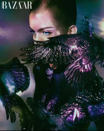 Fashion shot of a model wearing a dark dress adorned with bird-like embellishments, with a vibrant background, as featured in Harper's Bazaar.