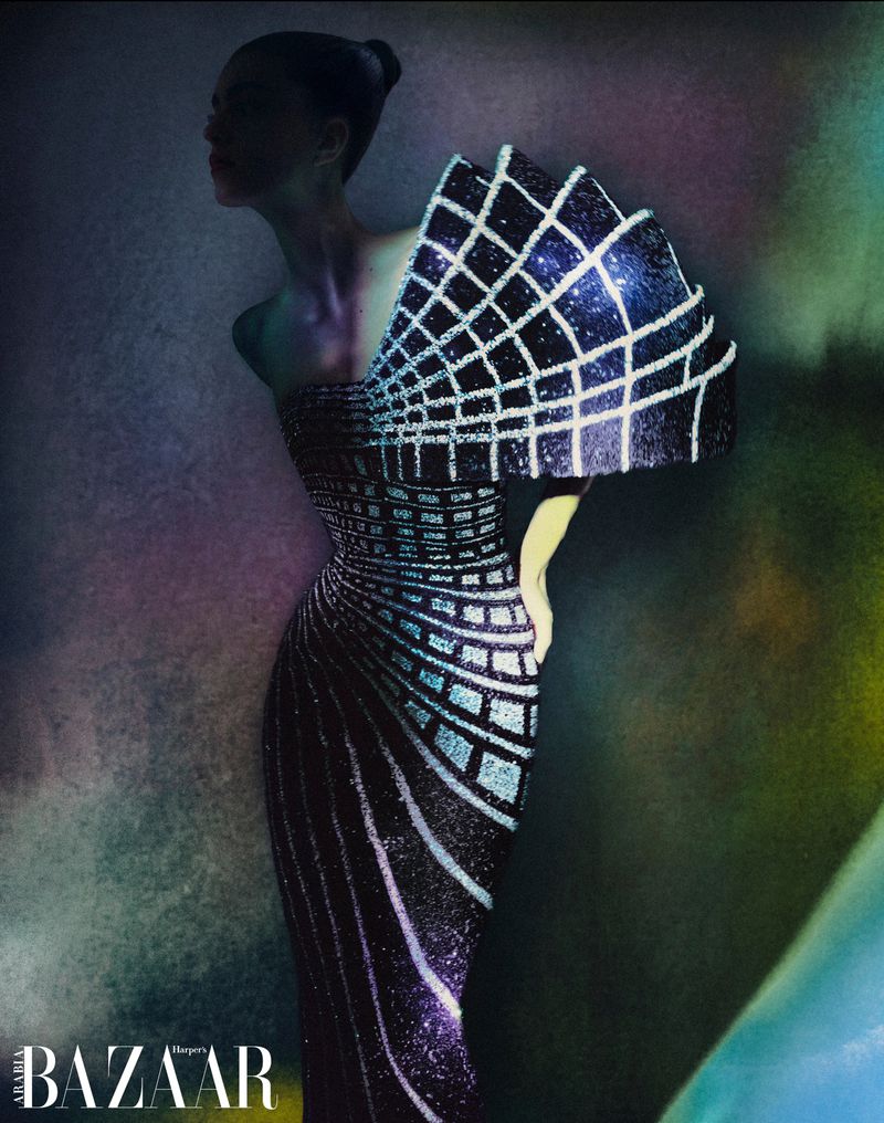 Woman in couture gown with grid pattern and dramatic shoulder detail posing in front of a textured multicolor backdrop, fashion editorial.