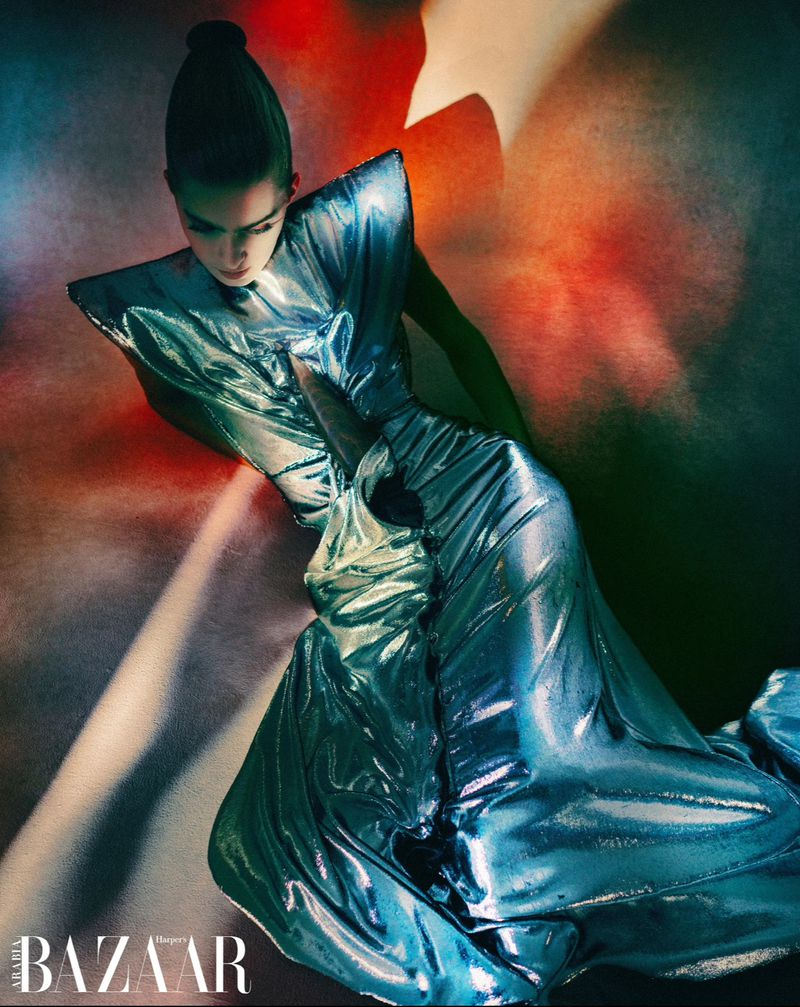 Fashionable woman poses in a shiny blue gown against a blurred background of red and green hues, embodying high-end fashion editorial.