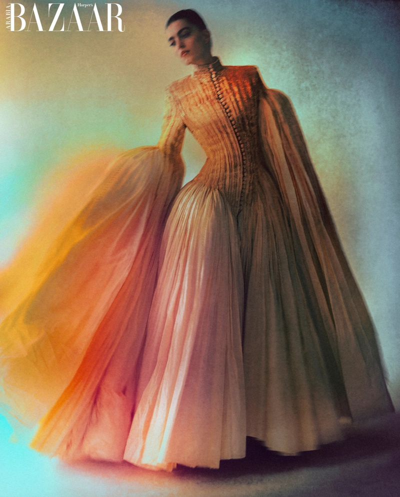 Fashion editorial shot of an adult female model in a long, light-gold gown with flowing sleeves, posing against a blurred background.