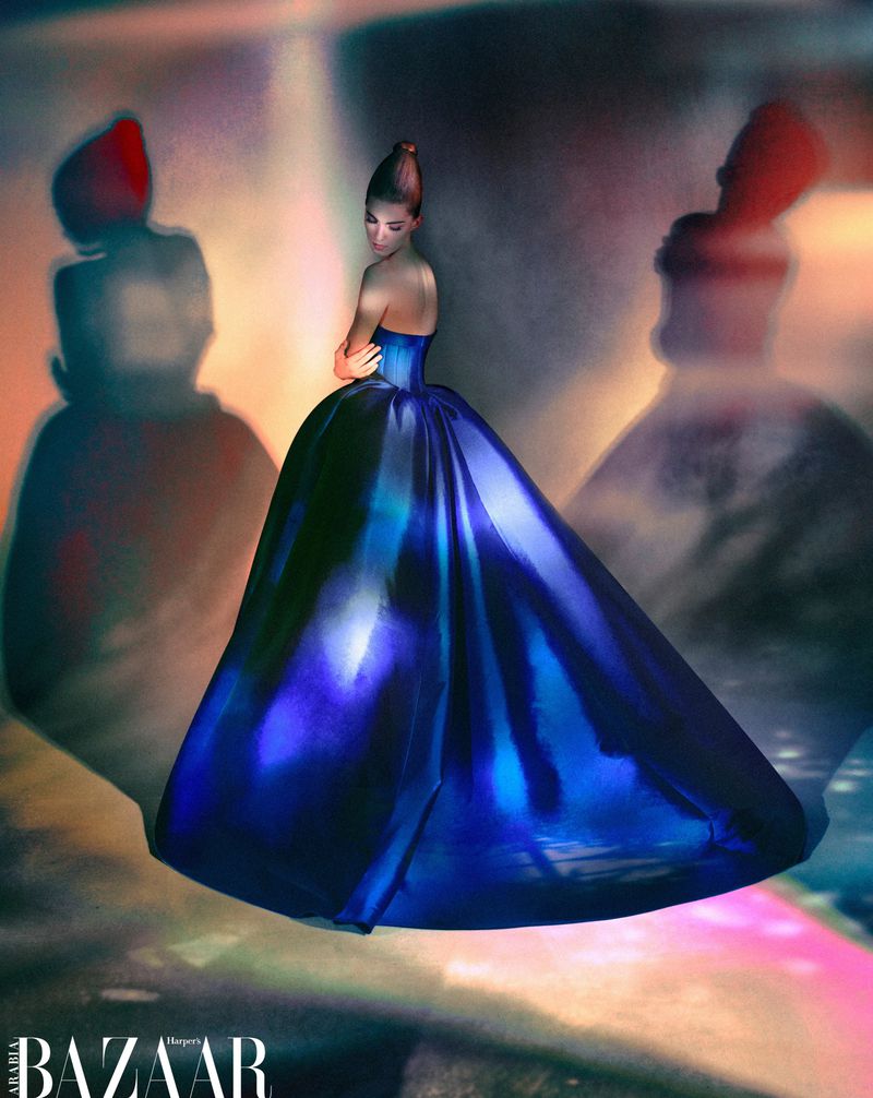 Woman in a formal strapless blue gown with a full skirt poses in a fashion editorial, with shadows of figures in the backdrop.