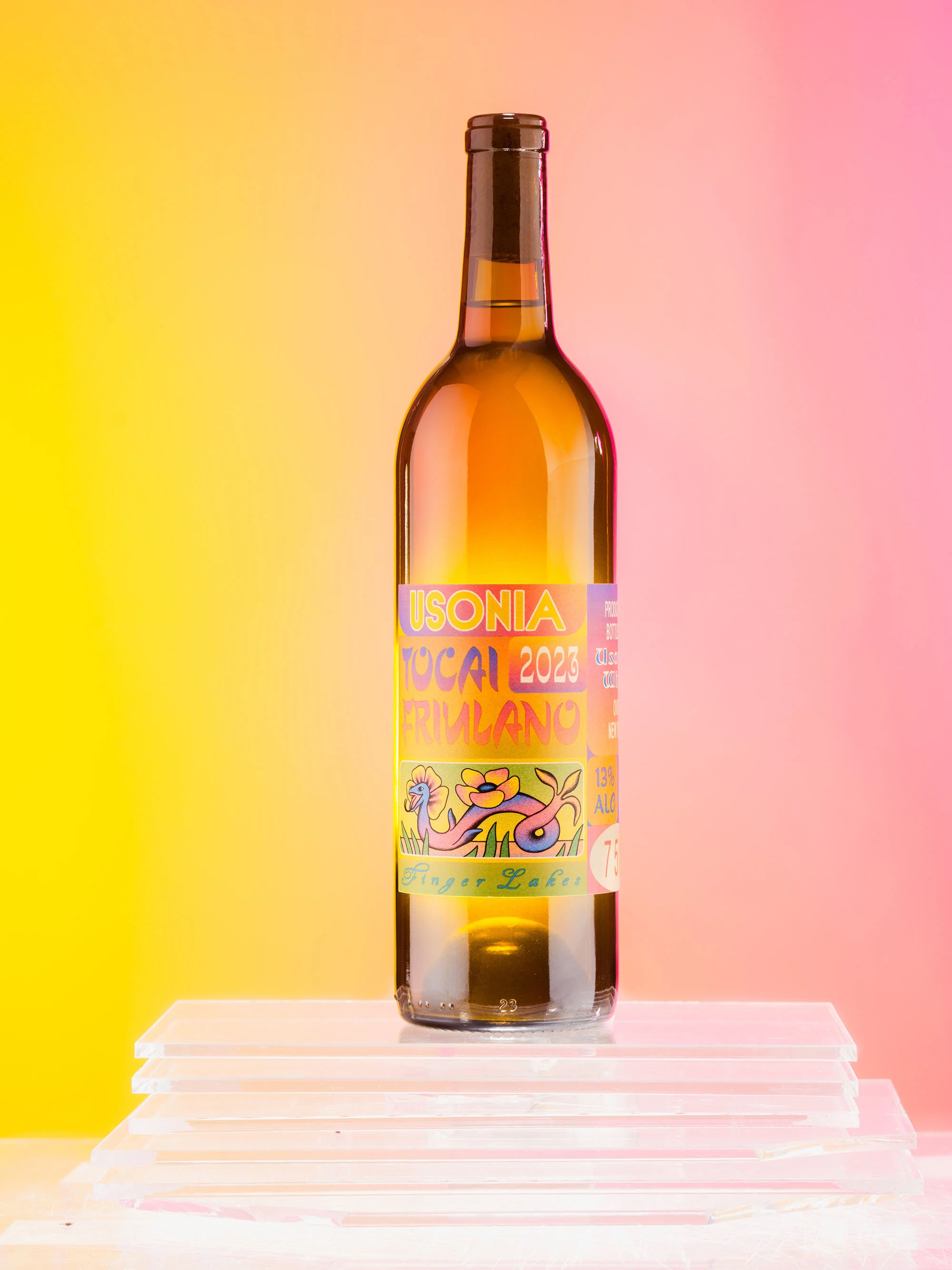Wine bottle with colorful label on a white platform against a yellow and pink gradient background. Product photography.
