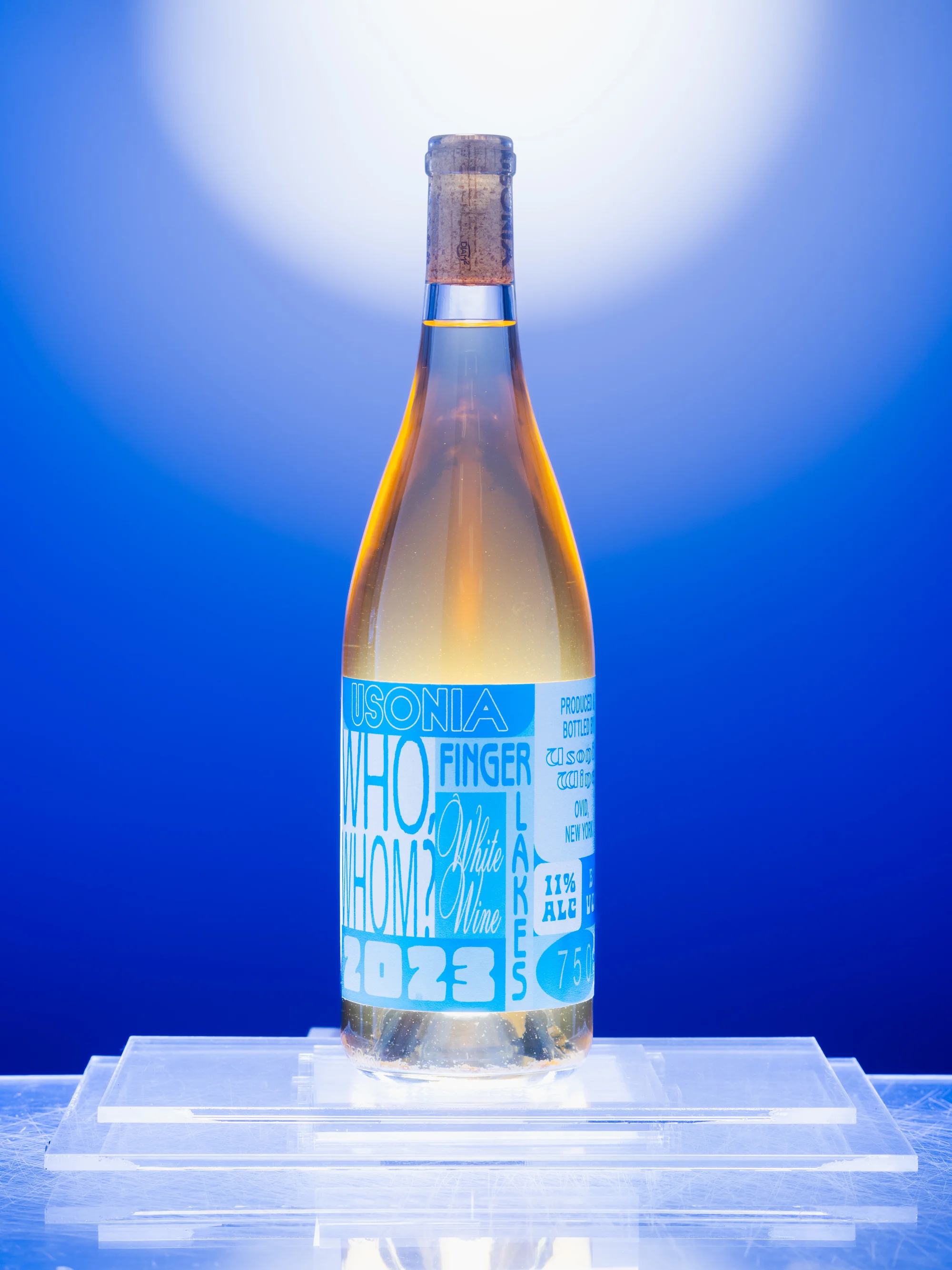 Bottle of Finger Lakes white wine with a cork top and blue label, placed on a clear acrylic stand against a soft blue background.