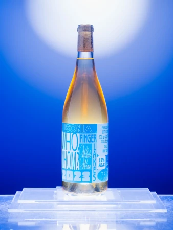 Bottle of Finger Lakes white wine with a cork top and blue label, placed on a clear acrylic stand against a soft blue background.