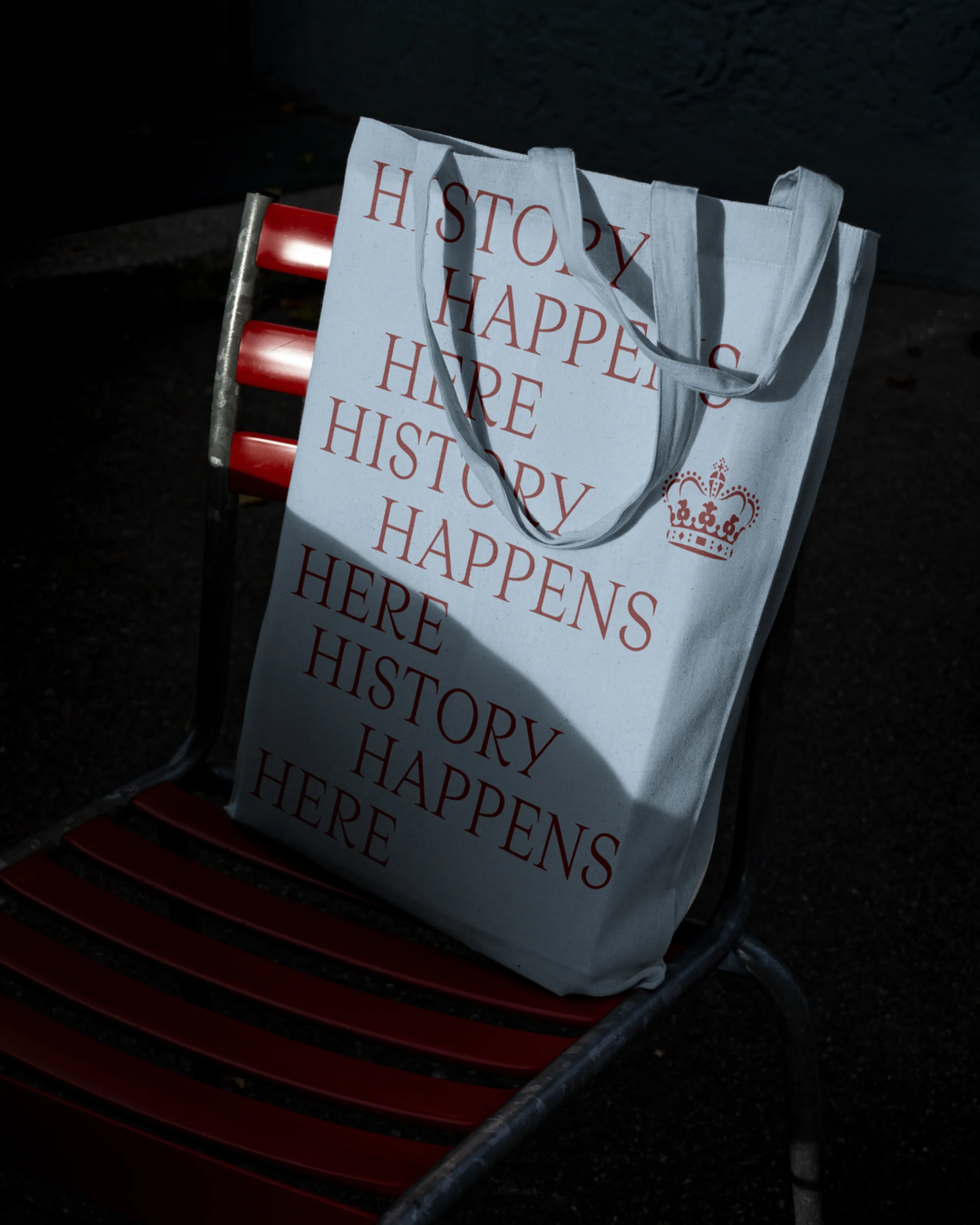Canvas tote bag with 'History Happens Here' text sitting on a red metal chair in a shadowed outdoor setting.