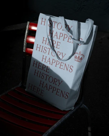 Canvas tote bag with 'History Happens Here' text sitting on a red metal chair in a shadowed outdoor setting.