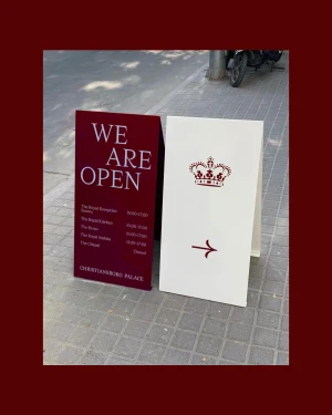 Two A-frame sidewalk signs for Christiansborg Palace, one burgundy with white text and one white with a red crown and arrow.