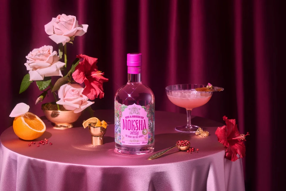 Bottle of rose and hibiscus gin with pink cocktail, floral arrangement, orange slice, and decorative spoon on pink tablecloth.