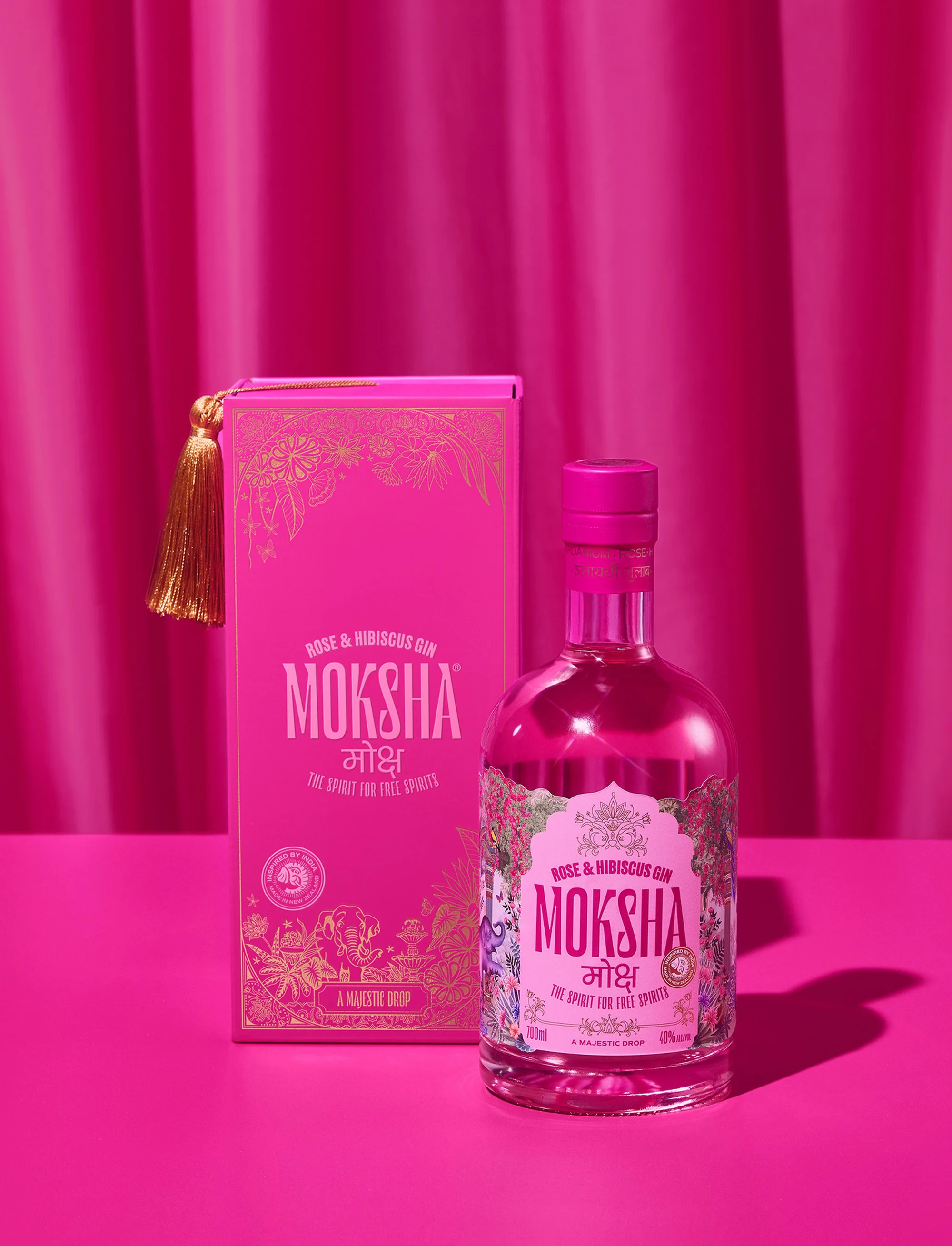 Rose and hibiscus gin bottle and matching box sit on a pink surface against a bright pink curtain backdrop in a studio setting.