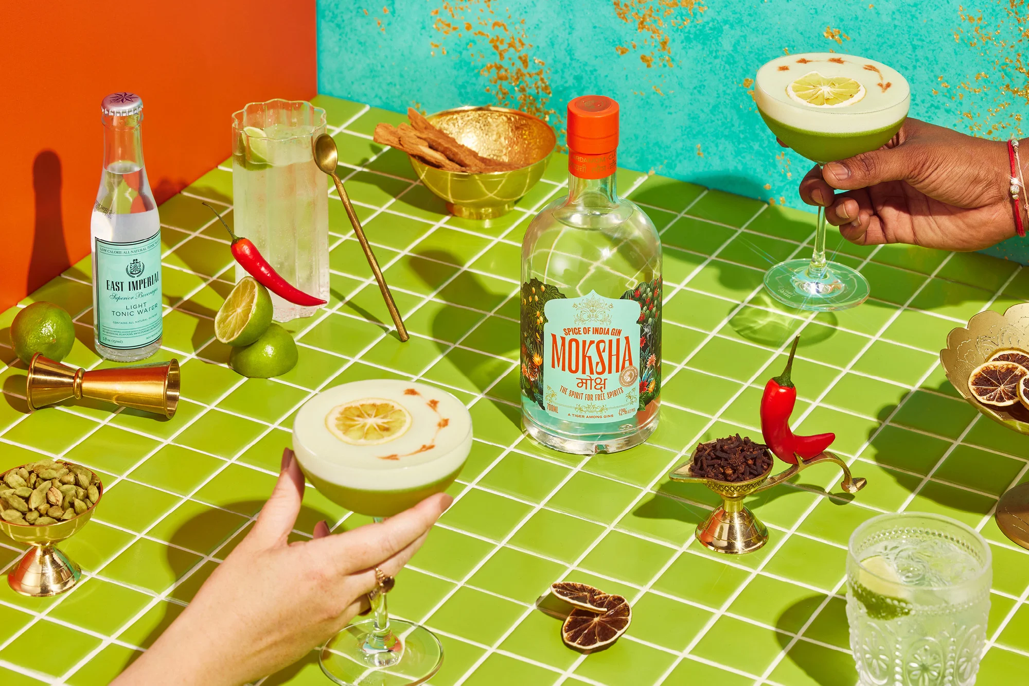 Still life of cocktail glasses, liquor bottle, tonic water, limes, and other items on a green-tiled table against a colorful background.