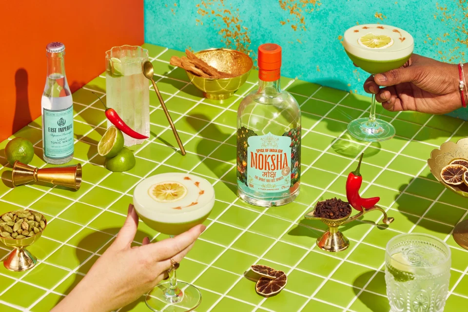 Still life of cocktail glasses, liquor bottle, tonic water, limes, and other items on a green-tiled table against a colorful background.
