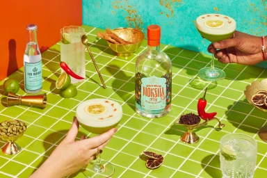 Moksha gin bottle surrounded by cocktails, tonic water, fresh limes, red chilis, and spices on a green tiled table.